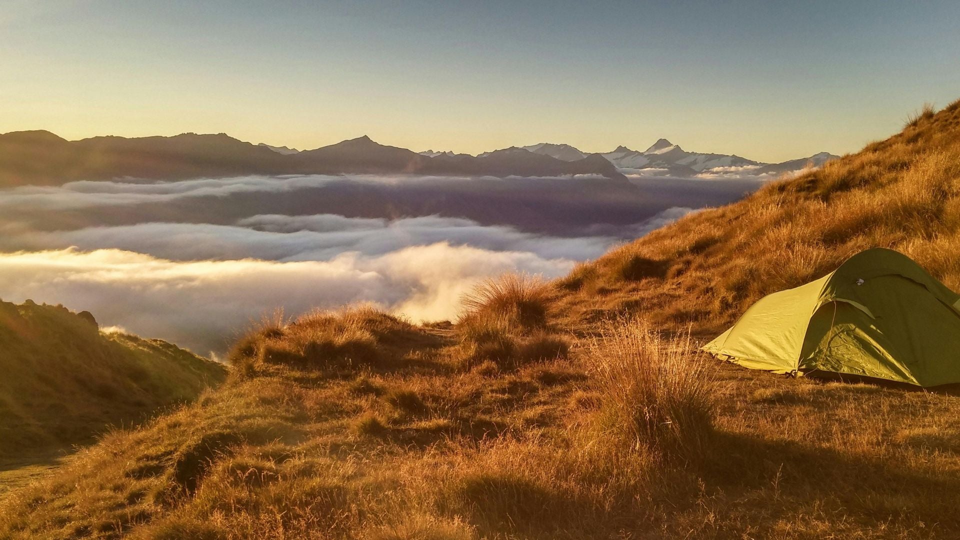 Green tent on a grassy hill with a mountainous landscape and clouds below at sunset.